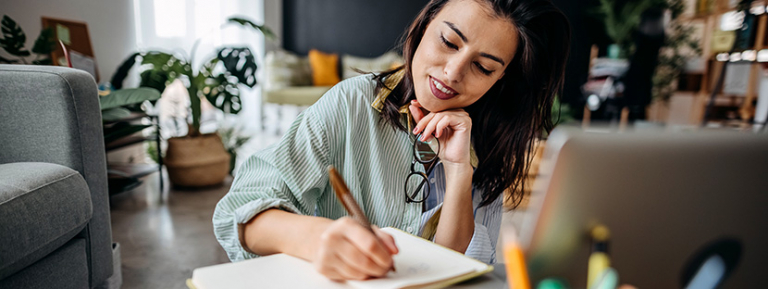 Young woman writing notes in a notebook, surrounded by home comfort.