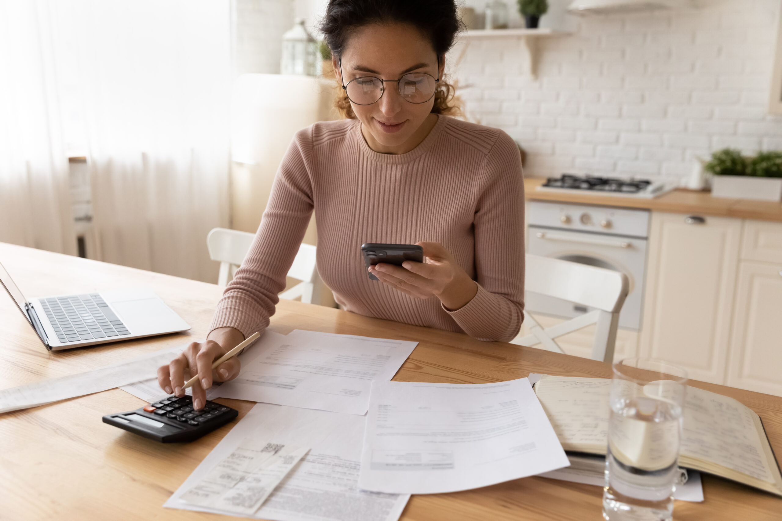 A woman with curly hair and glasses smiles while looking at her smartphone and adding up budget numbers on a calculator.