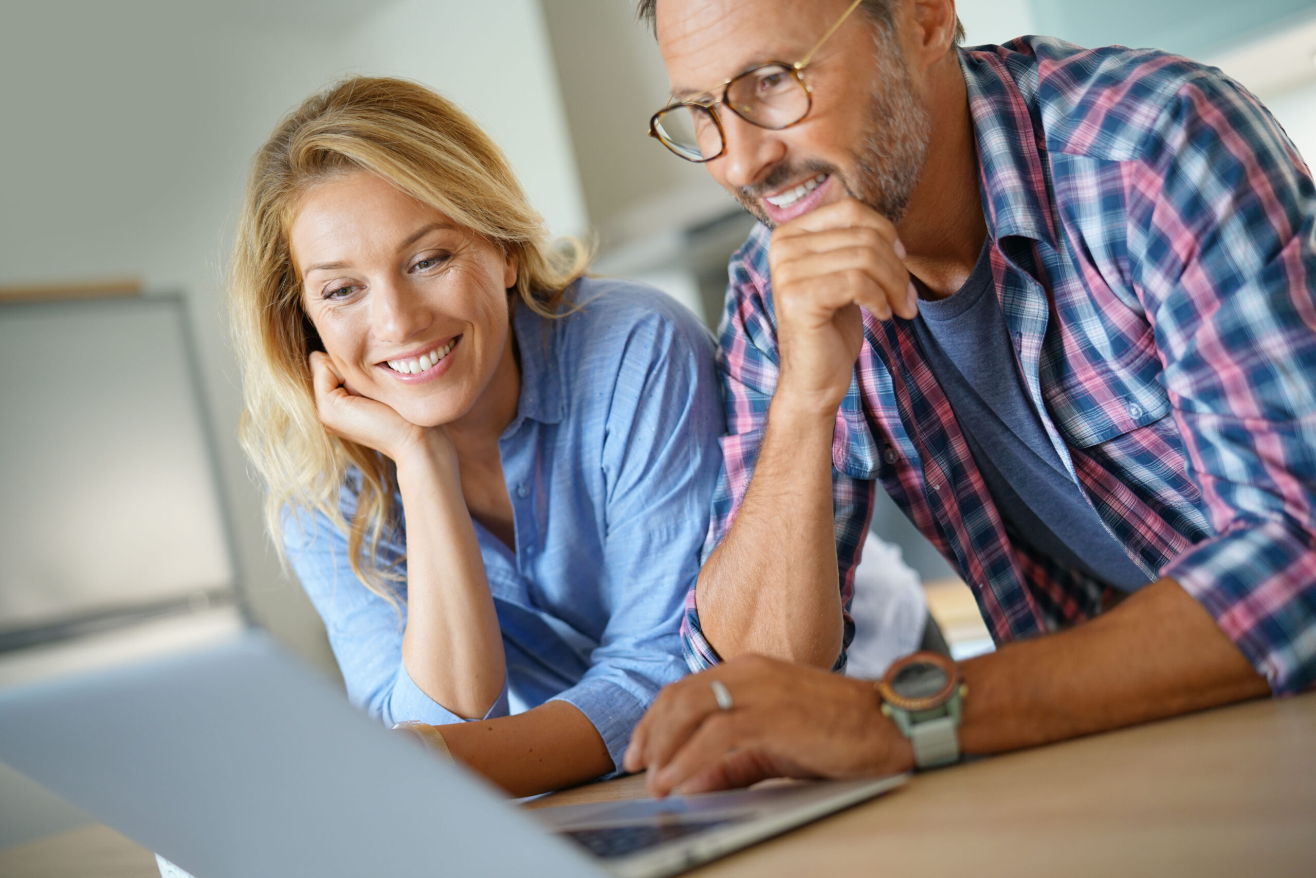 A smiling man and woman are seated at their kitchen table with an open laptop, looking at a mortgage payment estimate.